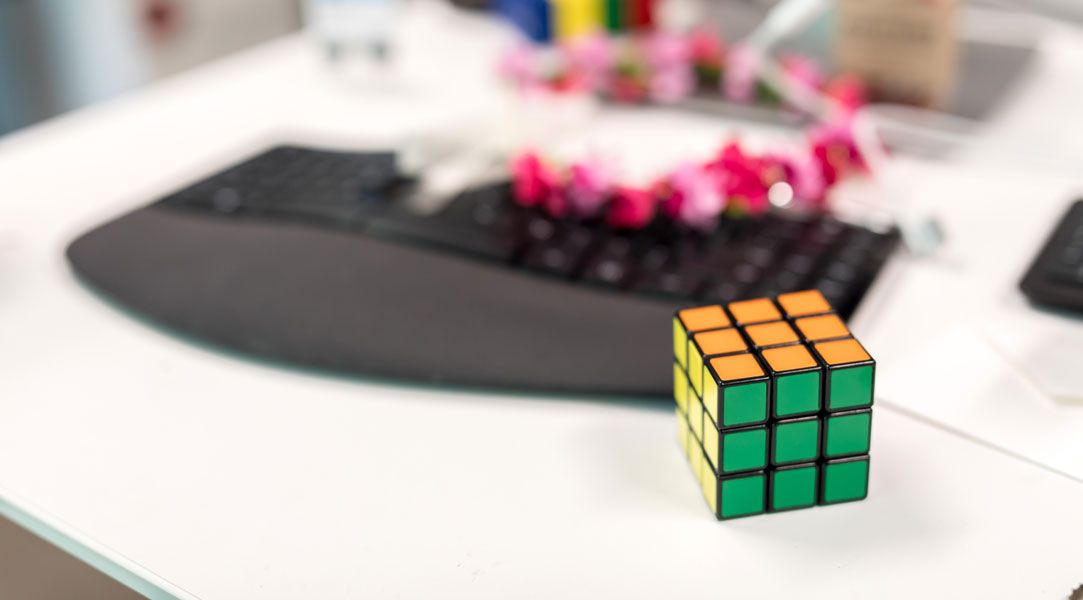 Nahaufnahme von Rubiks Würfel auf weißem Schreibtisch. Schwarze Tastatur, rosa Blumenketten und andere unscharfe Elemente, die im Hintergrund sichtbar sind.